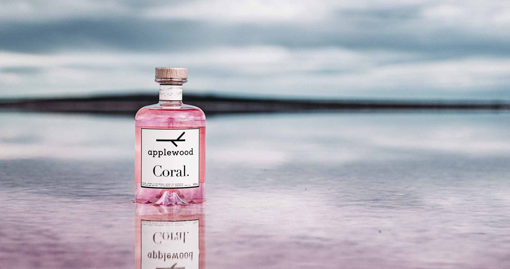 Applewood Coral pink gin on a beach. There is a blue sky and pink bottle with a beautiful reflection